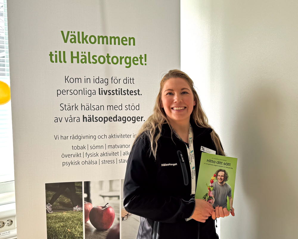 Hälsopedaog, ung kvinna med bok i handen står och ser in i kameran. Svarta kläder, långt blont hår. Affisch med hälsotorget bakom.