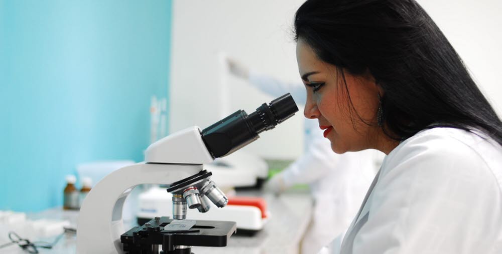 Kvinna i en vit laboratorierock använder ett mikroskop i ett laboratorium, med en suddig bakgrund där labbutrustning och en annan person syns.