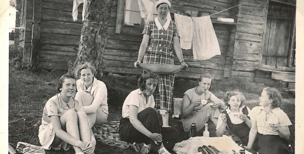 Ett svartvitt foto av en grupp människor som sitter och fikar på gräset.