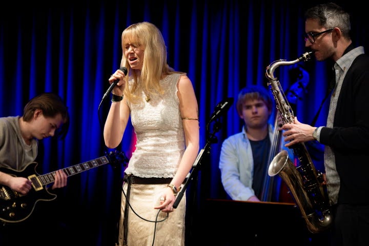 Musiker uppträder på scen med sång, gitarr, kontrabas och saxofon.